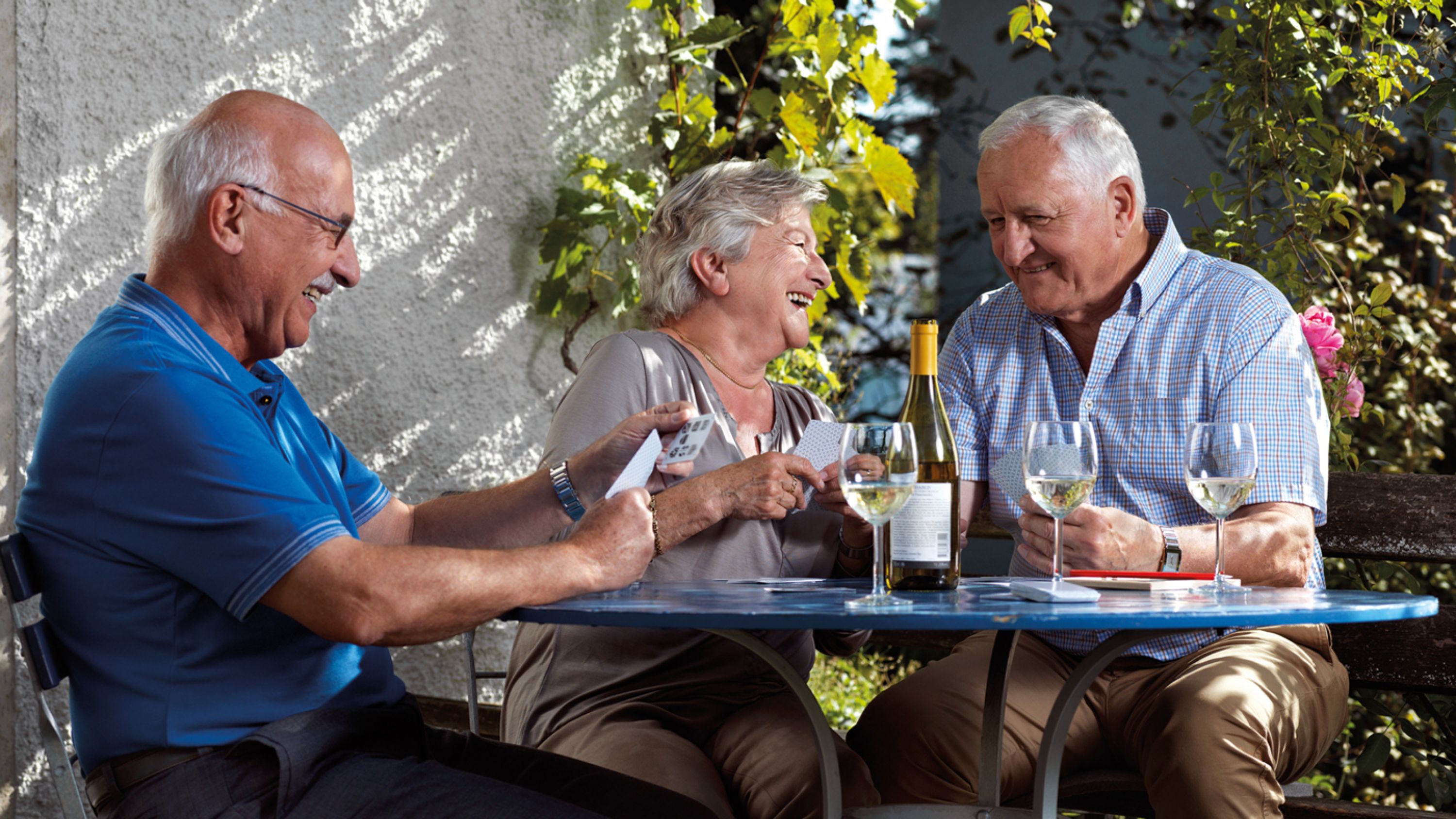 Trois personnes âgées sont assises à table, un verre de vin à la main, et rient
