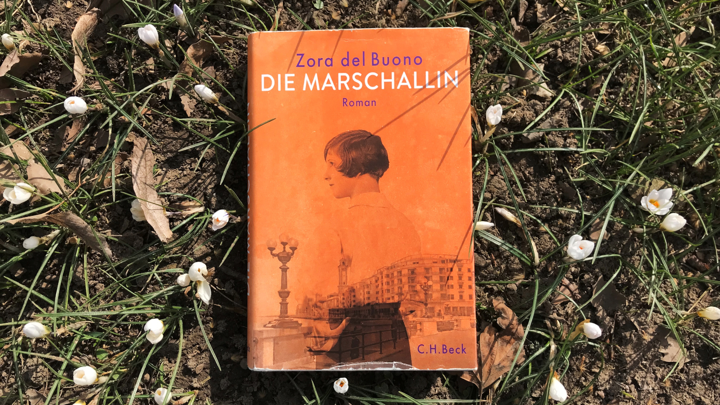 Buch "Die Marschallin" von Zora del Buono zwischen Schneeglöckchen liegend. Das orange Buchcover zeigt übereinandergelegt ein Bild einer jungen Frau und Häuser an einem Platz.