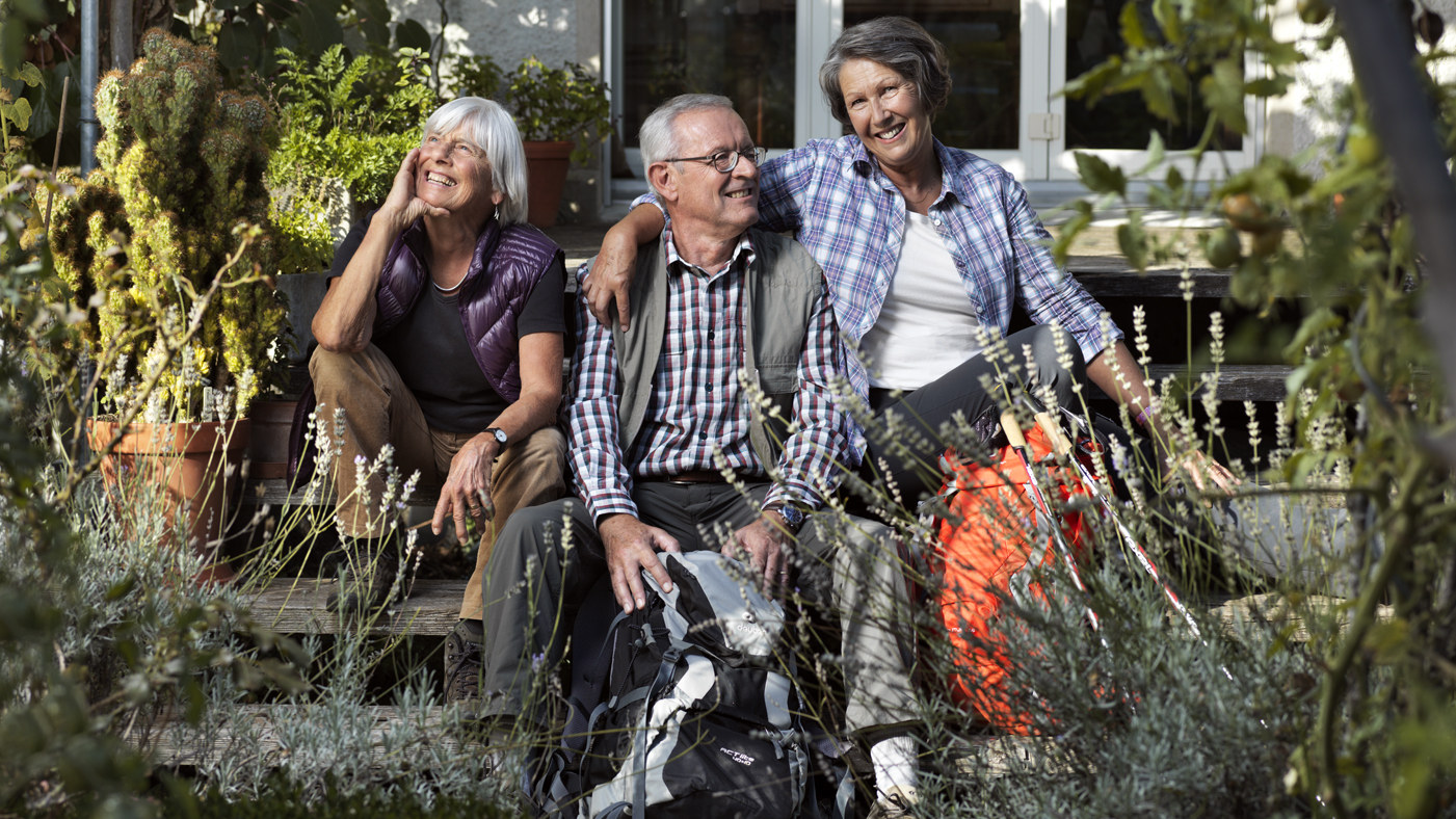 Zwei Seniorinnen und ein Senior mit Wanderausrüstung vor einem Haus am sitzen.